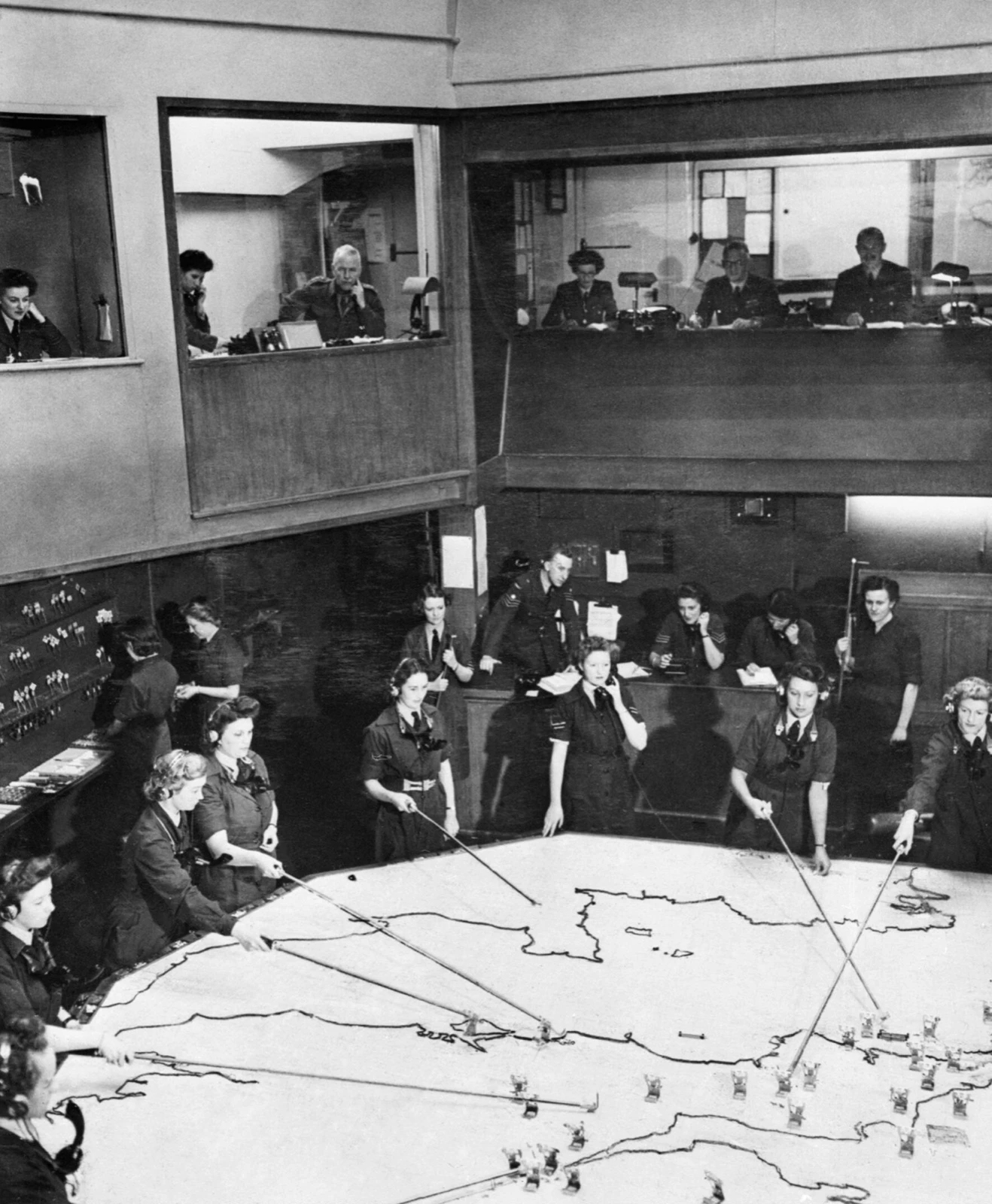 WAAF plotters moving aircraft markers across a giant map table with rakes while controllers watch from a gallery above