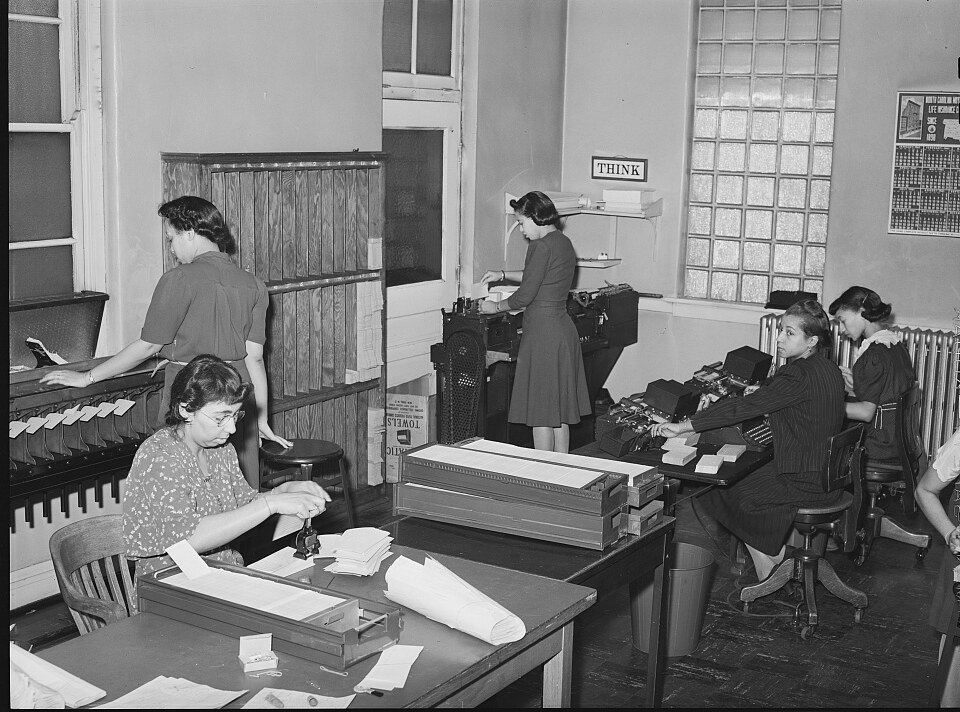 The office force of a Chicago insurance company in 1941: a clerk stamping paperwork at her desk, a colleague working a card file, and three women at adding machines and a typewriter