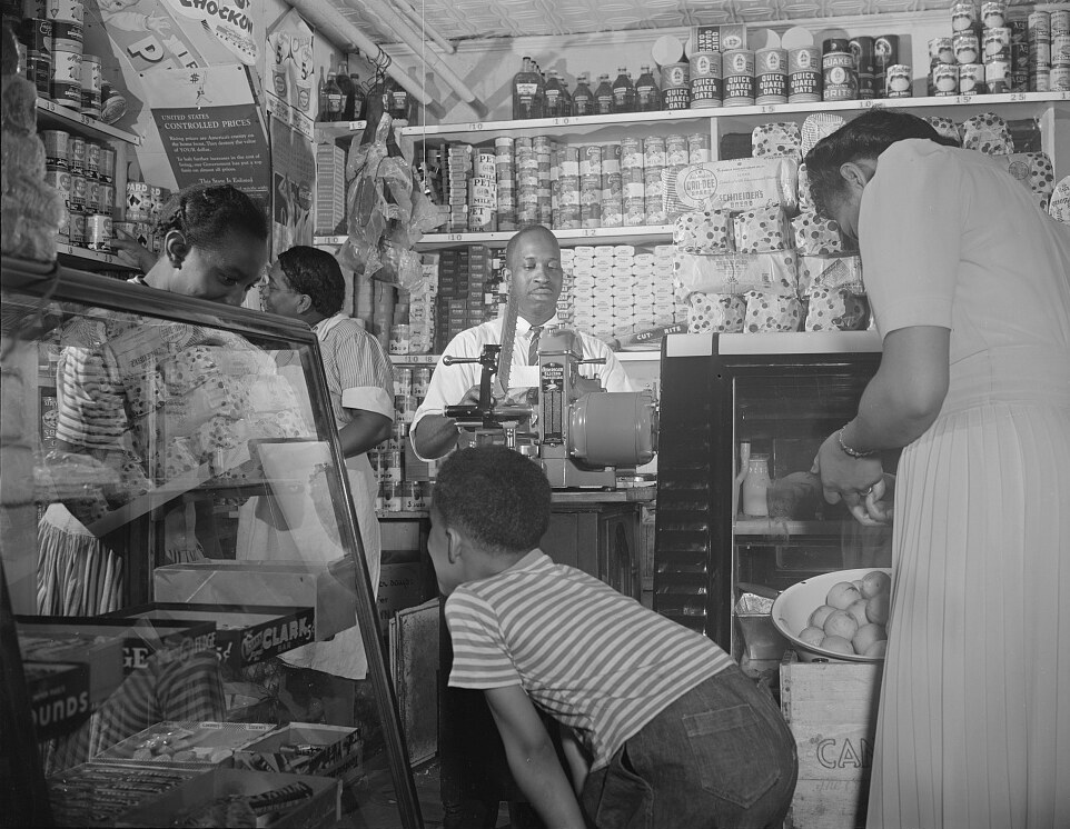 A Washington, D.C. grocery store on a Saturday afternoon in 1942: the owner working the slicer behind the counter, shelves stocked and price-tagged floor to ceiling, customers at the cold case and the candy cabinet